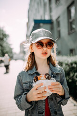 little girl in a denim jacket, silver cap and sunglasses eating street food on a sunny city street