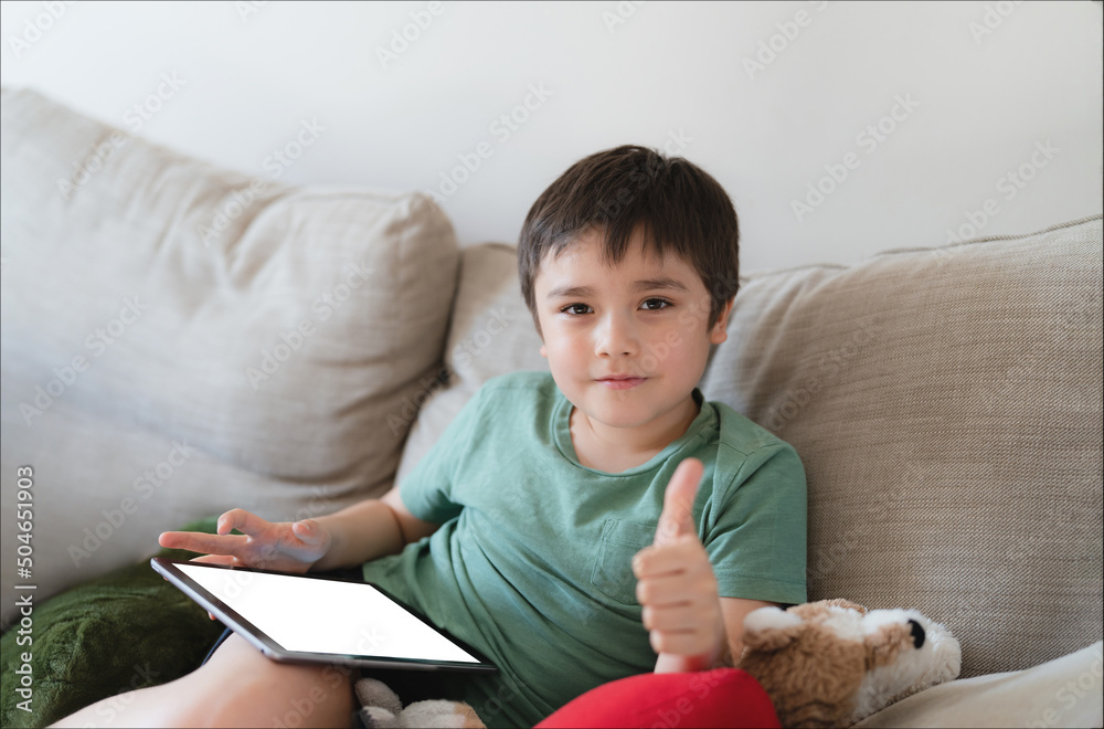 Wall mural Young boy using tablet playing game on internet with friend, Homeschooling Kid doing homework online by digital pad at home,Child sitting on bed holding touch pad maock up.