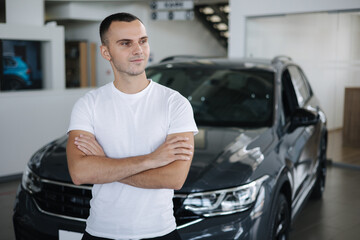 Portrait of handsome man standing by car in car shoowroom. Man buying new car. Male in white t-shirt. Happy person