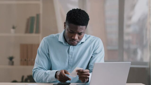 Young African focused man bank employee bookkeeper hold payment bill manage personal budget use calculator calculates expenses costs credit balance keeps accounting savings sitting at home office desk