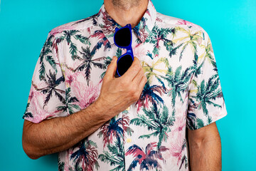 Man in a Hawaiian shirt holding blue colored glasses, with a green background. 