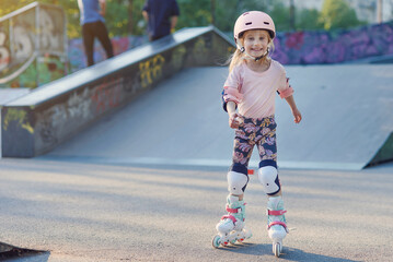 Little beautiful girl on roller skates in a helmet and protection of hands and feet in a skate park. Cheerful preschool girl skates on a sunny day.