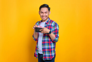Cheerful young Asian man in plaid shirt showing wallet with cash money isolated on yellow background