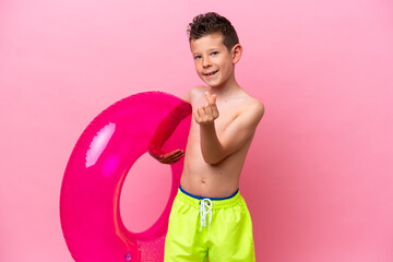 Little caucasian boy holding a inflatable donut isolated on pink background making money gesture © luismolinero