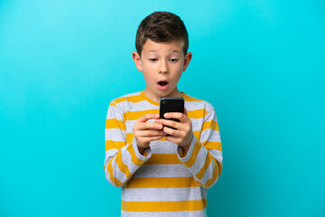 Little boy isolated on blue background looking at the camera while using the mobile with surprised expression