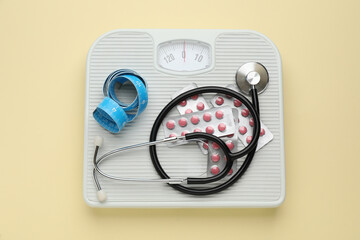 Scales with weight loss pills, measuring tape, and stethoscope on beige background, top view