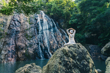 A young woman in white clothes and a turban sits on a rock near a waterfall, practices yoga and meditates