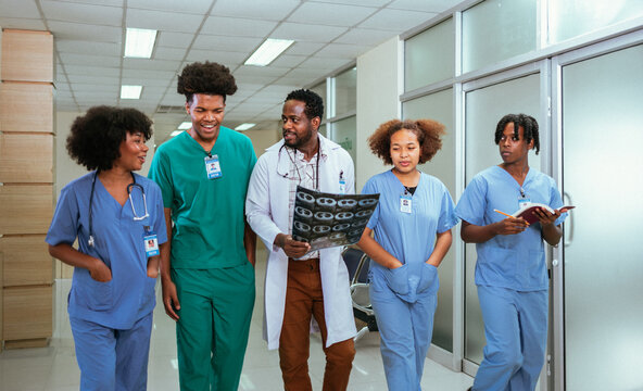 Group of medical students walking in a hallway at medical university. Focus is on African American male student.African doctors discussing MRI scan film on a hospital corridor in background.