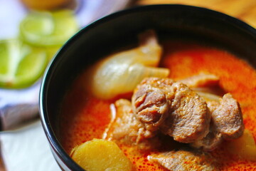 Massaman curry in black crockery served with boiled potatoes and chili on a brown wooden table.
