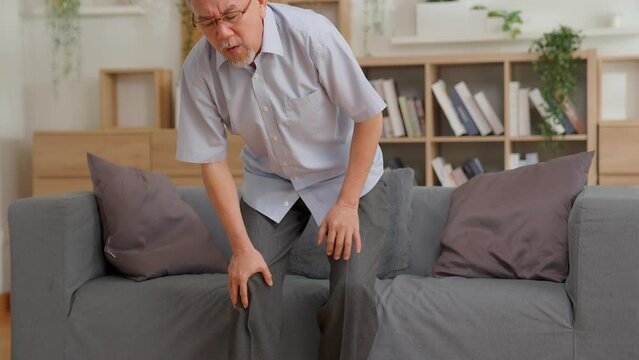 old Asian senior man feel knee joint pain on couch at home, ache, hurt at knee while standing and sitting at home, elderly knee pain osteoarthritis concept