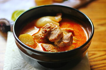 Massaman curry in black crockery served with boiled potatoes and chili on a brown wooden table.