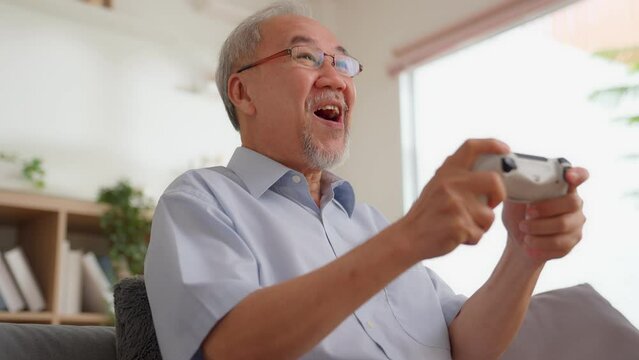 Asian senior man playing game on couch at home. Old people have fun using joysticks to control. A hobby was pro player gamer after retirement. Excited, exciting, celebrating, happy to win, gamer zone.
