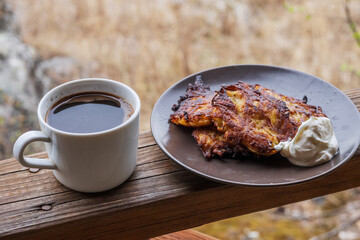 A plate of potato pancakes with sour cream and a mug of hot coffee stand on the wooden railing of...