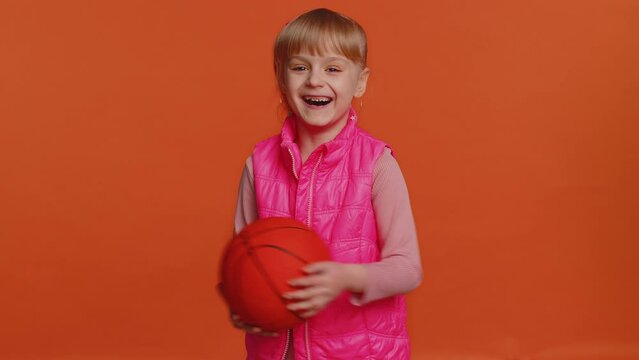 Girl sportsman basketball fan holding ball looking at camera, training dribbling. Workout sport motivation lifestyle concept. Young little blonde child kid isolated alone on orange studio background