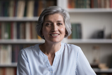 Positive happy older business teacher woman video call head shot portrait. Friendly smiling grey haired lady looking at camera, posing with library bookshelves in background