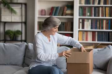 Happy elderly grey haired customer woman unpacking parcel at home, receiving purchase from internet...