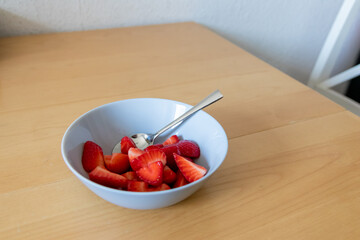 Many cut strawberries in bowl are ready to eat with silver spoon or as delicious finger food and healthy snack with red fruit and vitamins on wooden kitchen table for sustainable nutrition and health