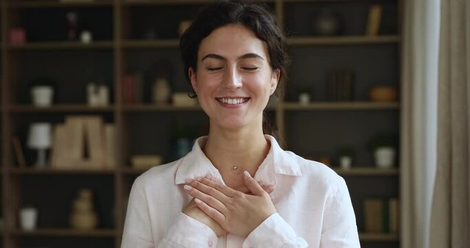 Head shot kind sincere young woman pose alone indoor put folded palms on chest looks grateful, feeling candid emotions smiles with eyes closed showing gesture of appreciation, thanking fate concept