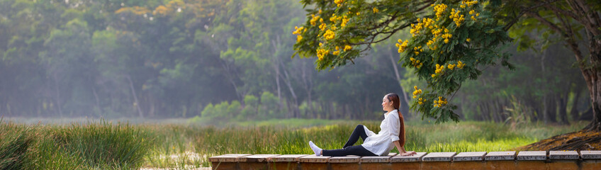 Asian woman sitting on the edge of dock with peaceful natural park during summer with yellow flower...