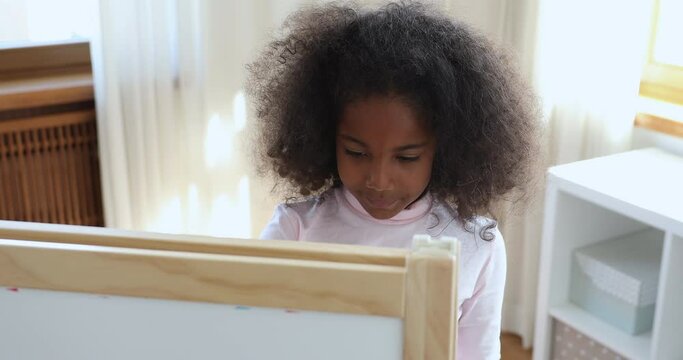 Serious African little curly-haired girl drawing on white board close up. Cute preschool child spend free time take part in art class standing alone in modern nursery room. Hobby, development concept