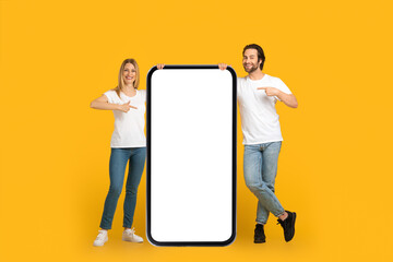 Satisfied millennial european couple in white t-shirts point fingers at huge phone with empty screen