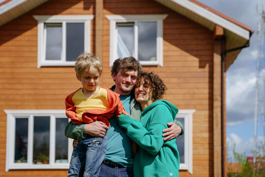 Happy Family: Dad Mom And Their Son Hug, Stand In Front Of Their House, Smile And Look At The Camera