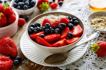 Healthy breakfast bowl, porridge with the addition of fresh berries; strawberries, blueberries and raspberries poured with honey in a ceramic bowl on a rustic table.