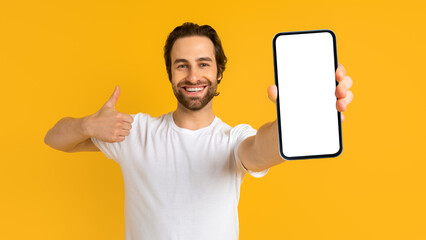 Glad young european man with stubble in white t-shirt show smartphone and thumb up with blank...