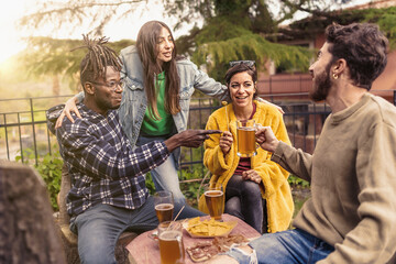 Multi ethnic roup of young friends enjoying talking and toasting beers sitting at the garden pub -...