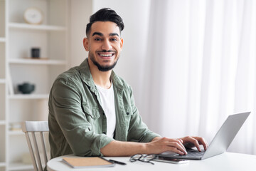 Smiling young islamic man manager with beard typing on laptop in home office interior, free space