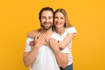 Cheerful millennial european female hugging from behind male with stubble in white t-shirts looking...