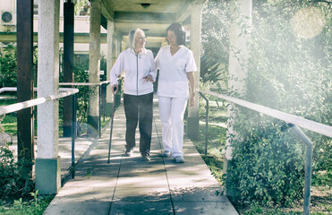 Asian doctor helping elderly retired woman with walker stick in the hospital yard. Rehabilitation concept