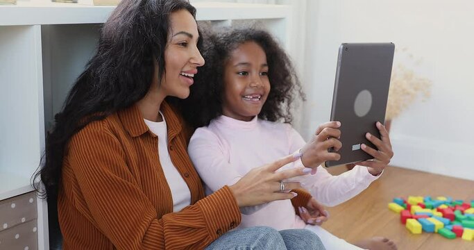 Close up happy little girl have fun use digital tablet make video call spend free time on internet at home with young African mother, young generation and modern tech overuse, fun, kid growth concept