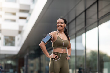 Young black woman in sportswear listening to music, using smartphone and fitness tracker during jogging on city street