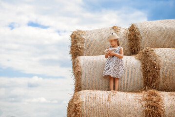 little girl in a straw hat  is standing on hay bales against the backdrop of clouds © Olena