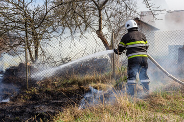 spring fire, burning dry grass near buildings in the countryside. Firefighter extinguishes the...
