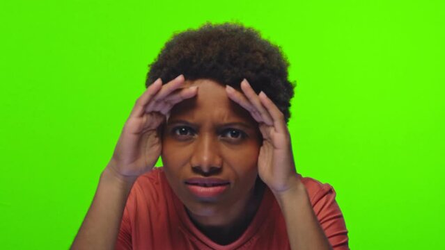 Young curious African American woman peering at screen and knocking on invisible glass greeting you rejoices and smiles dressed in casual T-shirt stands on green background in studio