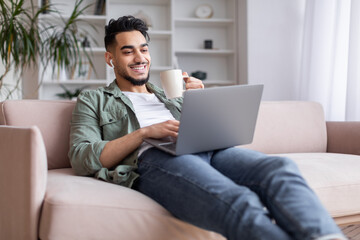 Glad millennial islamic guy with beard sits on sofa in wireless headphones, watch laptop, enjoy coffee and rest