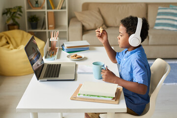 Side view of African-American boy in wireless headphones sitting at home desk and eating cookies while talking to online tutor