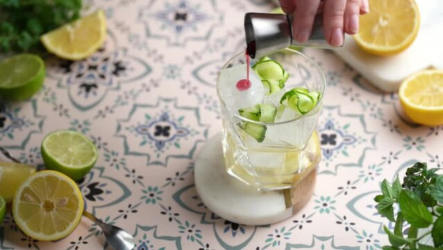 Cocktail lemonade preparation: female hand pouring red sirup from a measuring cupinto a glass with ice cubes on a multicolored portugal tiles background. Slow motion