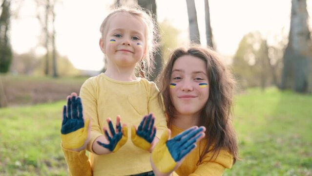 Portrait of two smiling girls with flag of Ukraine on face and palms. Two sisters wave their hands at camera.