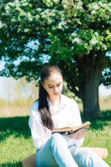 girl in a white blouse and blue jeans. Sitting near a flowering tree and reading a book.