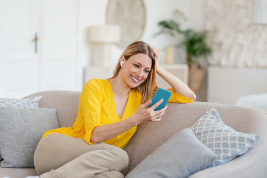 Cheerful young blonde european lady in yellow clothes with wireless headphones looks at phone, sits on sofa