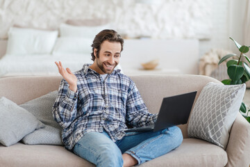 Obraz premium Satisfied millennial caucasian man with stubble sits on sofa waving hand and looking at laptop