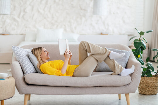 Happy pretty young blond european woman in yellow clothes lies on sofa and reads book