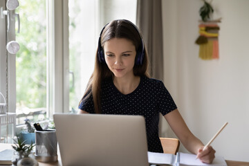 Positive focused student girl in headphones studying at home, attending online lesson, virtual class, learning workshop, watching webinar on laptop, writing notes. Internet education concept