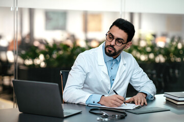 Serious attractive millennial arab guy doctor with beard in glasses, white coat looks at laptop, makes notes