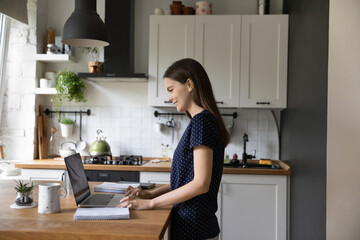 Happy pretty young renter girl using laptop in modern kitchen. Positive millennial gen Z student,...