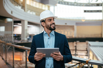 Cheerful millennial islamic guy engineer with beard in hard hat, suit checks project on tablet, looks at free space