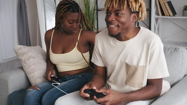 Couple home routine. Different hobby. Weekend leisure. Annoyed girl wearing headphones with music to reduce noise in room with excited guy playing video game on couch.